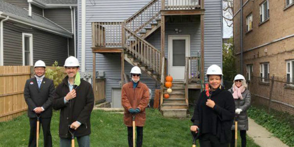 People wearing hard hats and coats pose with golden shovels on a lawn. A light-grey three story house is in the background.