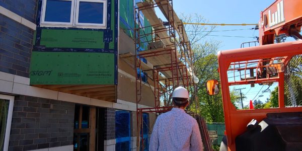 Man wearing a hard hat walks past on a construction site. A building without siding has scaffolding and a large construction vehicle is on the side.
