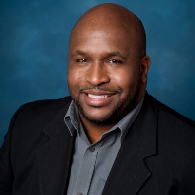 Bald black man with a blue-grey button shirt and a black suit jacket smiles in front of a blue background.