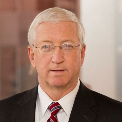 White man with white hair, wearing glasses, a white business shirt, red tie and dark suit.