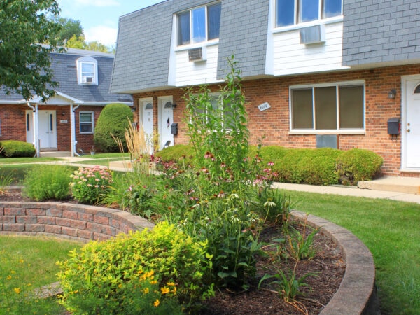 Flowers bloom in a curved raised garden bed made with light red brick. Two 2-story apartment buildings with red brick, black roofing tiles and white trim are in the background.