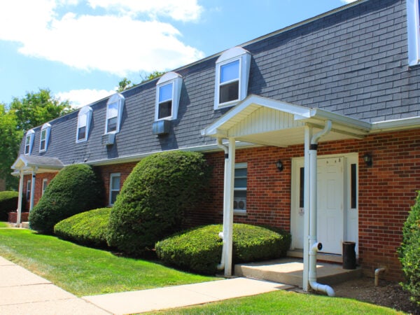 Two story apartment building with red brick, black roofing tile and white trim. Shrubs and grass are in the front yard.