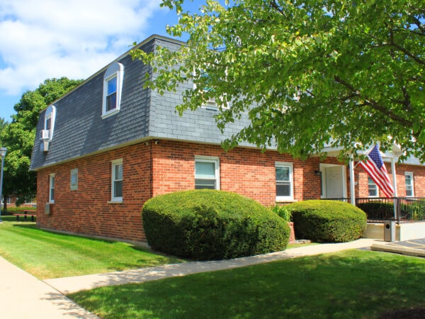 Two story apartment building with red brick, black roofing tile and white trim. A large tree, shrubs and grass are in the front yard.