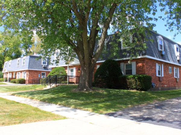 Two story apartment building with red brick, black roofing tile and white trim. A large tree, shrubs and grass are in the front yard.