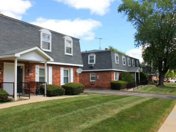 Two 2-story apartment buildings with red brick, black roofing tile and white trim. A large tree, shrubs and grass are in the front yard.