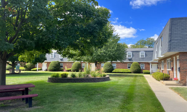 A large courtyard with green grass, raised flower beds and a large tree is lined by two-story apartment buildings with red brick, black roofing tiles, and white trim. A blue sky with fluffy white clouds caps the scene.