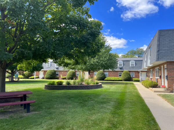 A large courtyard with green grass, raised flower beds and a large tree is lined by two-story apartment buildings with red brick, black roofing tiles, and white trim. A blue sky with fluffy white clouds caps the scene.