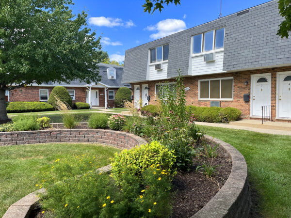 Flowers bloom in a curved raised garden bed made with light red brick. Two 2-story apartment buildings with red brick, black roofing tiles and white trim are in the background.