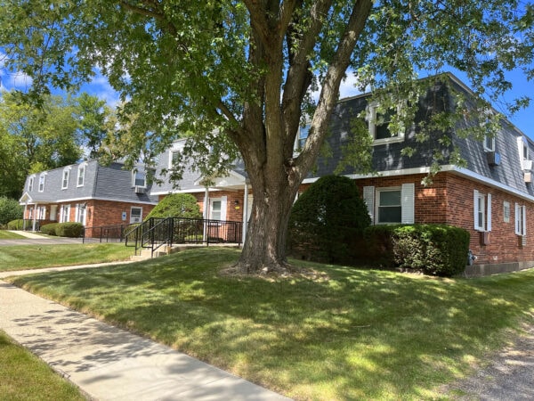 Two story apartment building with red brick, black roofing tile and white trim. A large tree, shrubs and grass are in the front yard.
