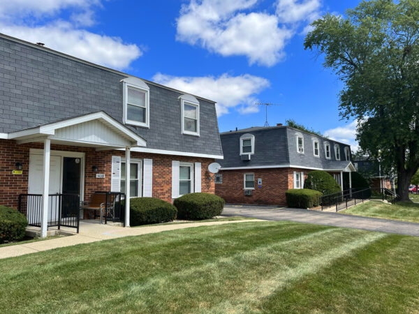 Two 2-story apartment buildings with red brick, black roofing tile and white trim. A large tree, shrubs and grass are in the front yard.