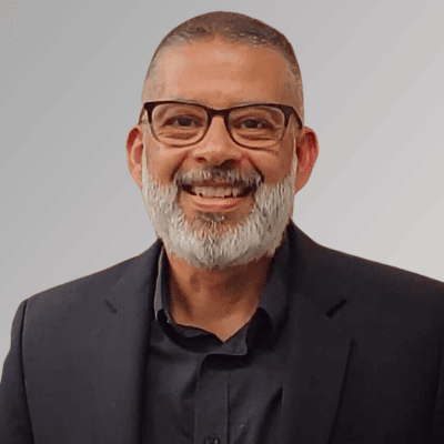 Man with short salt and pepper hair and beard, wearing glasses and a black button down shirt and black blazer.