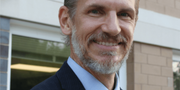 Man with salt and pepper short beard, wearing a light blue shirt, dark blue tie and suit, smiles in front of a tan brick building.