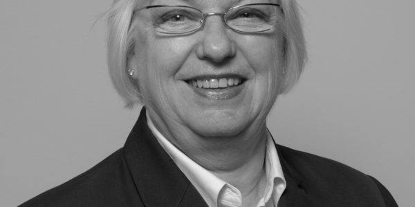 Black and white photo of a smiling woman with straight white hair wearing glasses, a white button down shirt, and a dark blazer.
