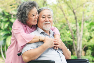 Elderly Asian man in a wheelchair and wearing a light blue shirt is hugged by an elderly Asian woman wearing a pink shirt.