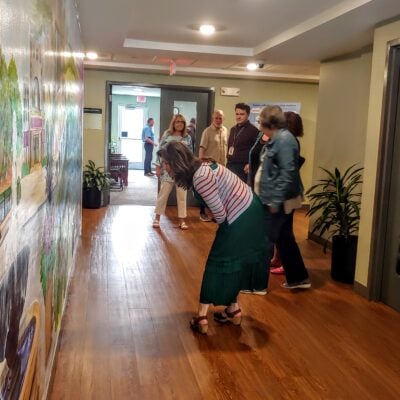 People stand in a hallway with a large mural on one side. A woman is bending over looking closely at the mural.