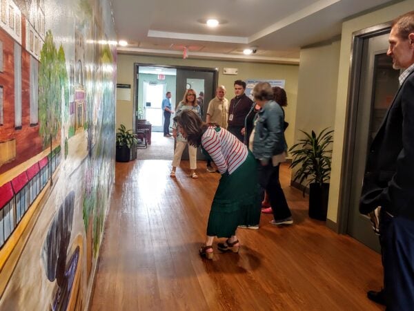 People stand in a hallway with a large mural on one side. A woman is bending over looking closely at the mural.