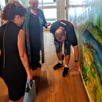 A man bends down, pointing a point of a mural on a wall, while three white women look on.