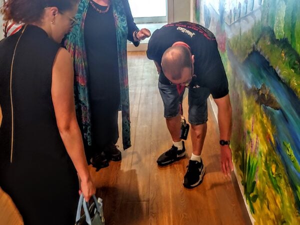 A man bends down, pointing a point of a mural on a wall, while three white women look on.