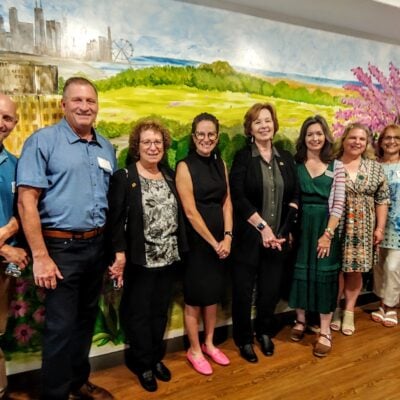 Group of professionally dressed people pose and smile in front of a large mural of green pastures and a tree with pink blossoms.