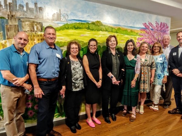Group of professionally dressed people pose and smile in front of a large mural of green pastures and a tree with pink blossoms.