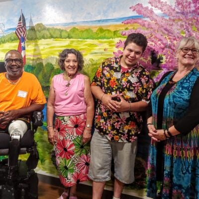 Four people smile in front of a wall with a painted mural of green pastures and a tree with pink blooms in the background. One black man is in a wheelchair. Two women and another man stand next to him.