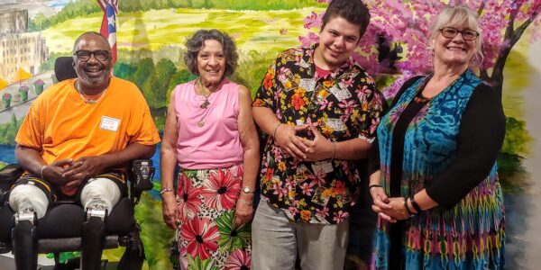 Four people smile in front of a wall with a painted mural of green pastures and a tree with pink blooms in the background. One black man is in a wheelchair. Two women and another man stand next to him.