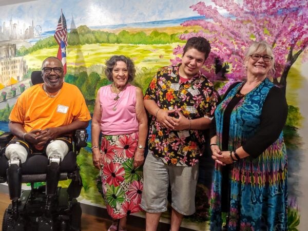 Four people smile in front of a wall with a painted mural of green pastures and a tree with pink blooms in the background. One black man is in a wheelchair. Two women and another man stand next to him.