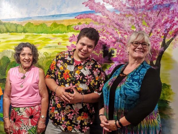 Four people smile in front of a wall with a painted mural of green pastures and a tree with pink blooms in the background. One black man is in a wheelchair. Two women and another man stand next to him.