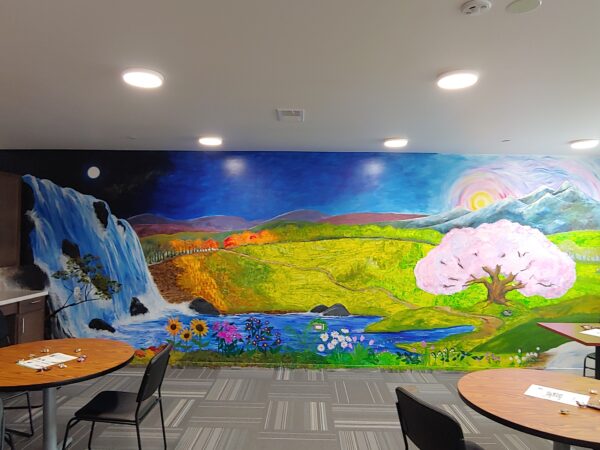 A large mural is painted on a wall. It has a landscape that includes mountains, green fields, a tree with pink blossoms, flowers, trees that have fall colors, a waterfall, a rising sun and a full moon. Round tables are in front of the mural with papers and small candies sprinkled on them.