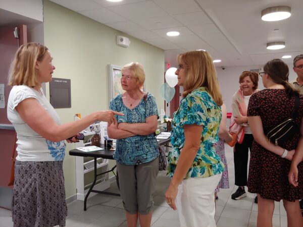 Three women are talking in a hallway while a group of people stand behind them talking to each other. There are balloons and a table with decorations and paper in the background.
