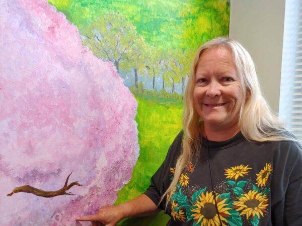 A woman with long blond hair and wearing a black t-shirt with sunflowers smiles as she points to a painted mural on a wall that includes a green meadow and a tree with pink blossoms. Painted into the pink blossoms is a small pink awareness ribbon for breast cancer. The woman is pointing to the painted ribbon.