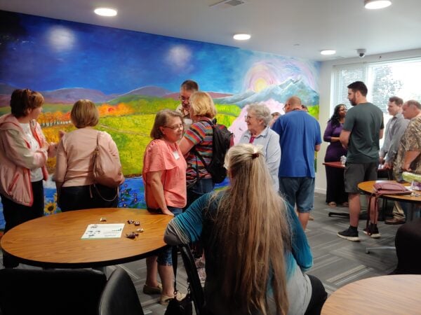People mingle as they stand in front of a large mural of a landscape with mountains, a rising sun, and green meadows. A woman with long grey-brown hair sits in front of two women who are talking with her.