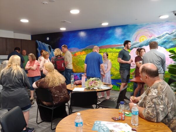 People mingling in front of a mural of a landscape. Some people are standing and others are sitting. Bottles of water, papers, and a bunch of flowers are on top of small round tables.