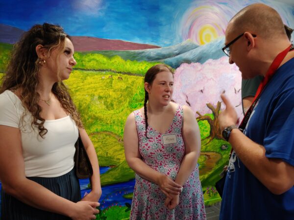 Two women talk with a man wearing a blue t-shirt as they stand in front of a mural of a landscape with green fields and a tree with pink blossoms.