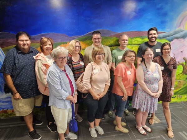 A group of women and men of various ages pose and smile in front of a wall mural that depicts a naturescape with a waterfall, mountains, and a tree with pink blossoms.