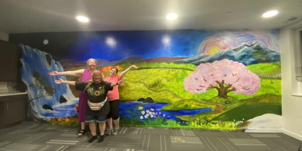 Three women with stand in front of a mural of a landscape with a large pink tree, green meadows, and a waterfall.