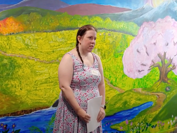 A woman with braided hair and wearing a summer dress stands in front of a mural of a landscape with a green meadow and a tree with pink blossoms. She is holding papers in her hands.
