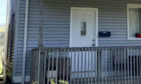 Front door of home with light blue-grey siding, a wooden porch, and small green shrubs.