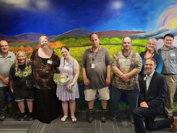 A group of women and men of various ages pose and smile in front of a wall mural that depicts a naturescape with a waterfall, mountains, and a tree with pink blossoms.
