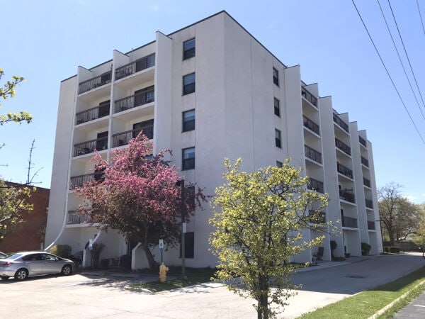 A six story white apartment building with balconies. A tree with pink blossoms, a smaller tree, and a yellow fire-hydrant are in front. A silver car isparked in the parking lot of the building that is framed by a white sky.