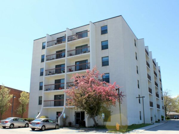 A six story white apartment building with balconies. A tree with pink blossoms and a yellow fire-hydrant are in front. Two silver cars are parked in the parking lot of the building that is framed by a white sky.