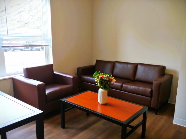 Living room with brown leather couch and chair. A coffee table with a warm wood top and black trim has a vase of fresh flowers. A window with half-opened blinds lets sunlight into the room with wood floors.