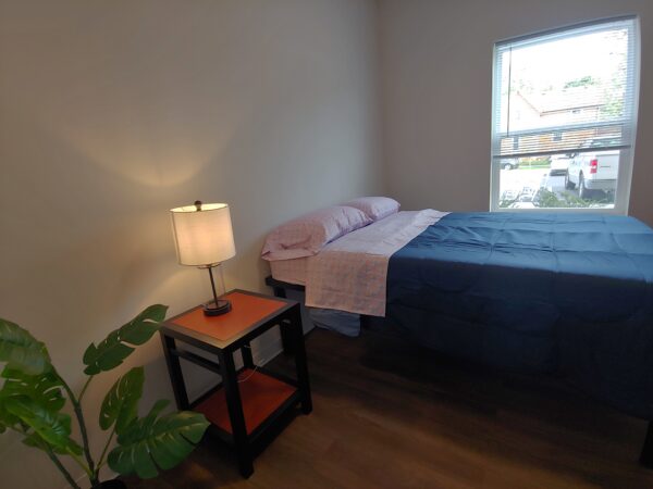 A bedroom with a double-sized bed with light sheets and a blue blanket. A lit lamp is on a small wooden side-table.