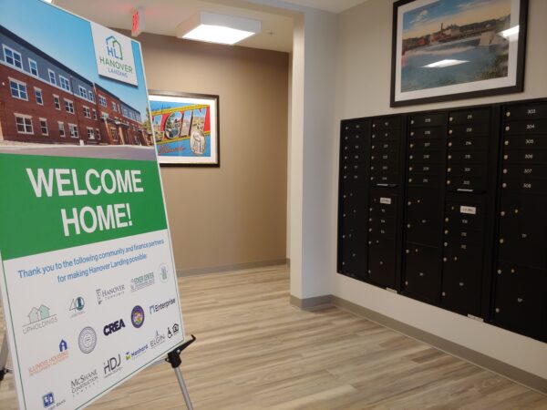 A welcome home sign stands in the main lobby with wooden laminate floors, a wall of mailboxes painted dark gray, and a light tan wall with artwork in the background.