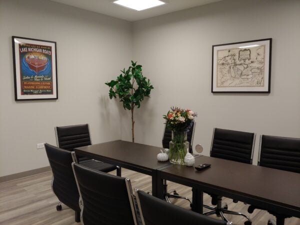 A conference room with a narrow dark brown table is surrounded by black swivel chairs. Artwork is on the walls and a large decorative tree stands in the corner.