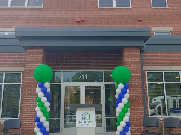 Balloon towers with white, green, and blue balloons are on either side of a main entrance to a red-brick building. A podium with the Hanover Landing logo stands in front.