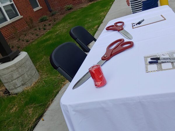 Two red-handled large scissors rest on a table covered with a white tablecloth. Papers and a roll of red ribbon are also on the table.