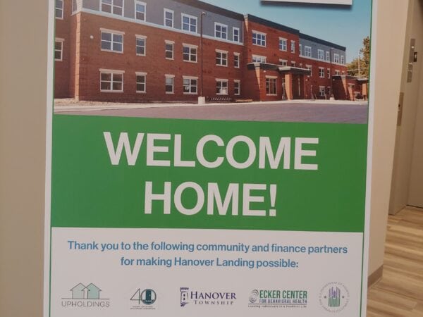 A large sign that says Welcome Home! in white text over a green background. A large picture of a red brick and light blue-grey siding apartment building is across the top. Along the bottom are a series of logos of the organizations that helped make the development a reality.