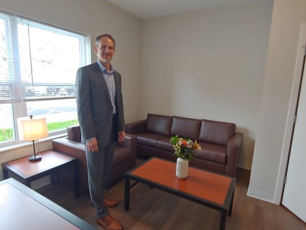 A tall, thin white man stands in a living room with a brown leather couch and matching chair. A wooden coffee table has a white vase with colored flowers. Light streams through the window behind the man who is wearing a grey suit and light button-down shirt.
