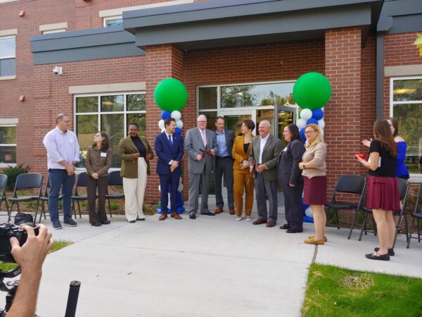 Men and women stand in pose in front of a red brick building. Large green balloons mark the sides of the main entrance behind the people.